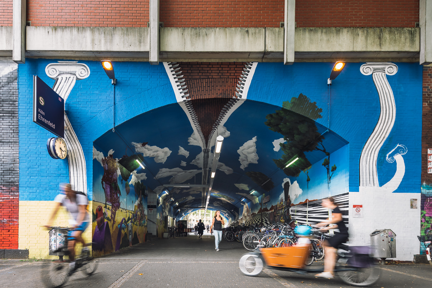 Bunte Graffiti-Unterführung am Bahnhof Köln-Ehrenfeld. Das Kunstwerk zeigt eine optische Illusion, bei der der Himmel durch einen Reißverschluss geöffnet wird. Seitlich sind verzerrte Säulen abgebildet. Mehrere Personen gehen oder fahren mit dem Fahrrad durch die Passage, daneben stehen zahlreiche geparkte Fahrräder.