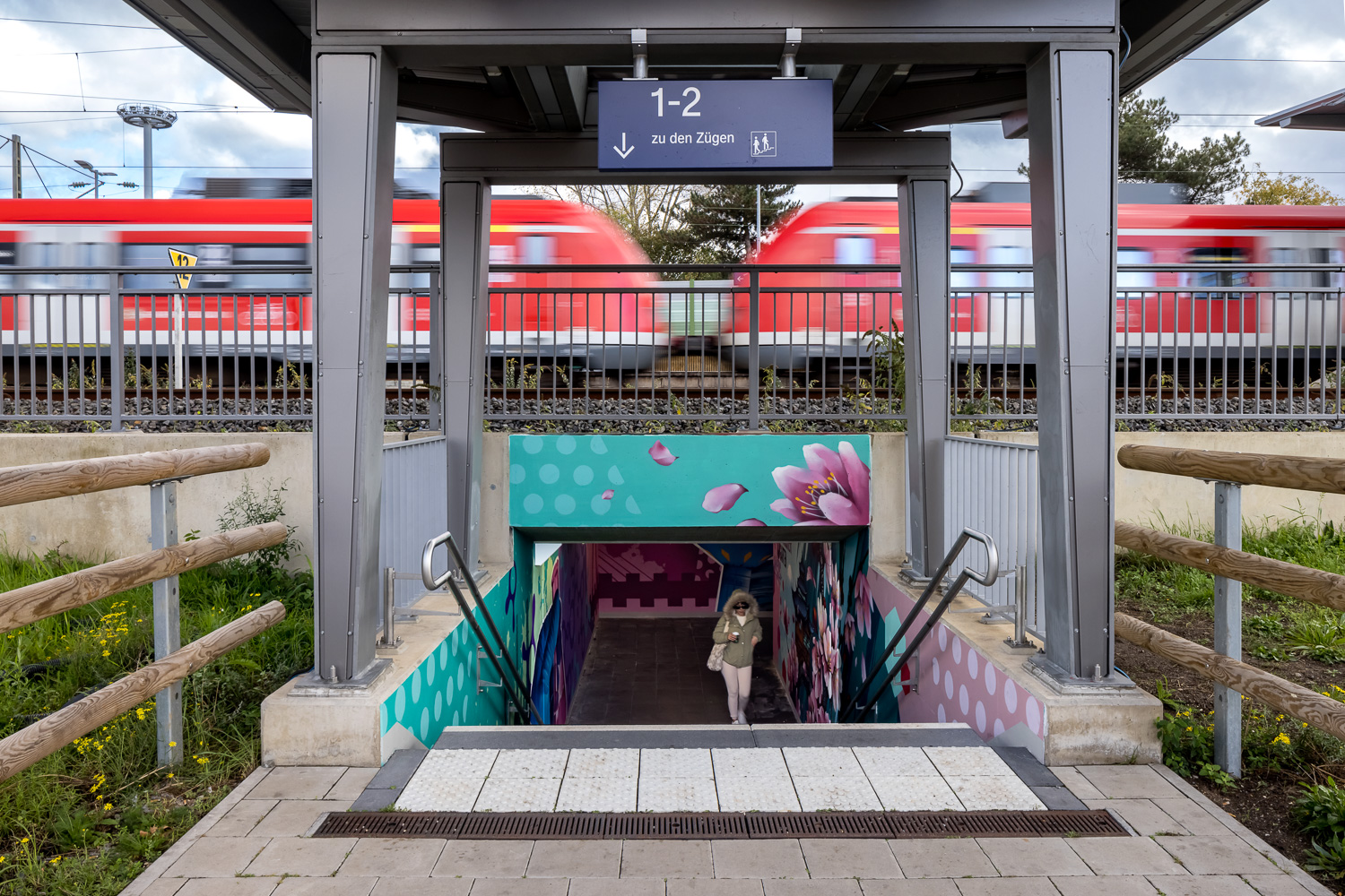 Eine Person geht eine Treppe von einem mit Wandmalereien verzierten S-Bahn-Durchgang in einem Bahnhof hinauf, während im Hintergrund zwei rote Züge vorbeifahren. Ein Schild über ihr zeigt die Bahnsteige 1-2 mit einem Pfeil nach unten an.