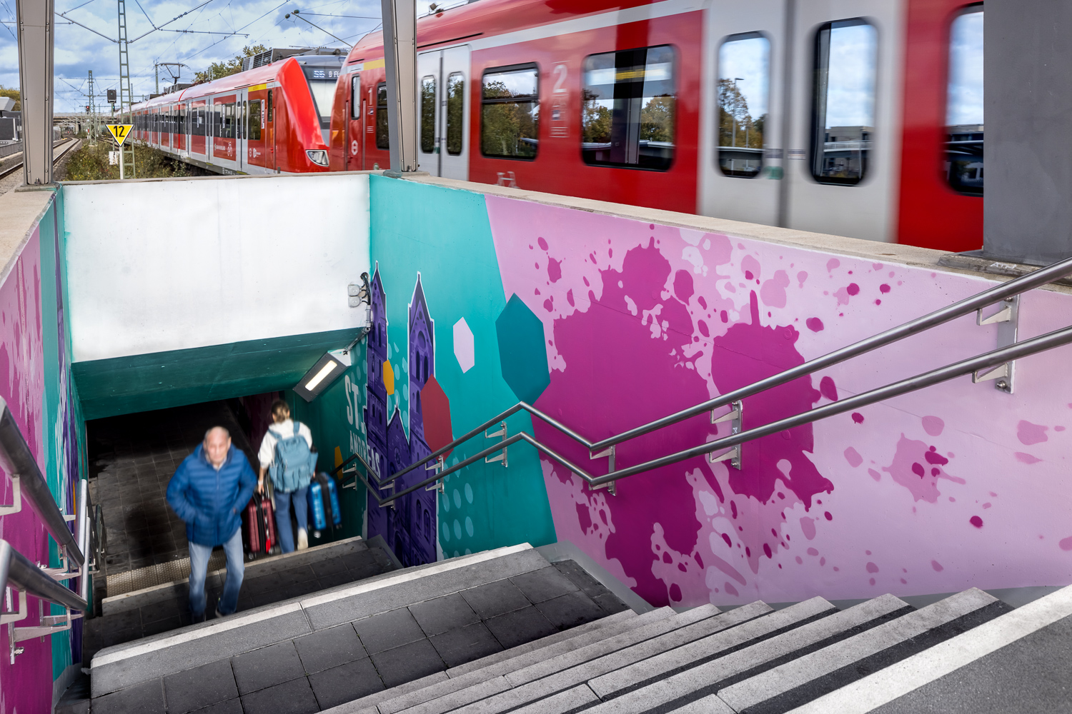 Ein Mann und ein Kind gehen eine Treppe von einer Unterführung mit buntem Graffiti hinauf, während oben auf den Gleisen ein roter Zug vorbeifährt. Die Szene spielt sich im Freien ab, und sowohl der Zug als auch die Treppe sind zu sehen.