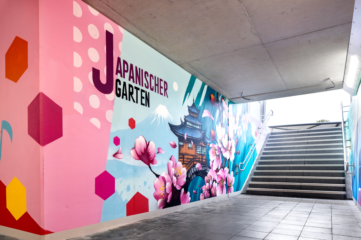 Ein bunt bemaltes Wandgemälde in einer Unterführung zeigt Kirschblüten, eine japanische Pagode, den Berg Fuji und den Schriftzug "Japanischer Garten" neben rosa Sechsecken und Punkten, während rechts eine Treppe nach oben führt.