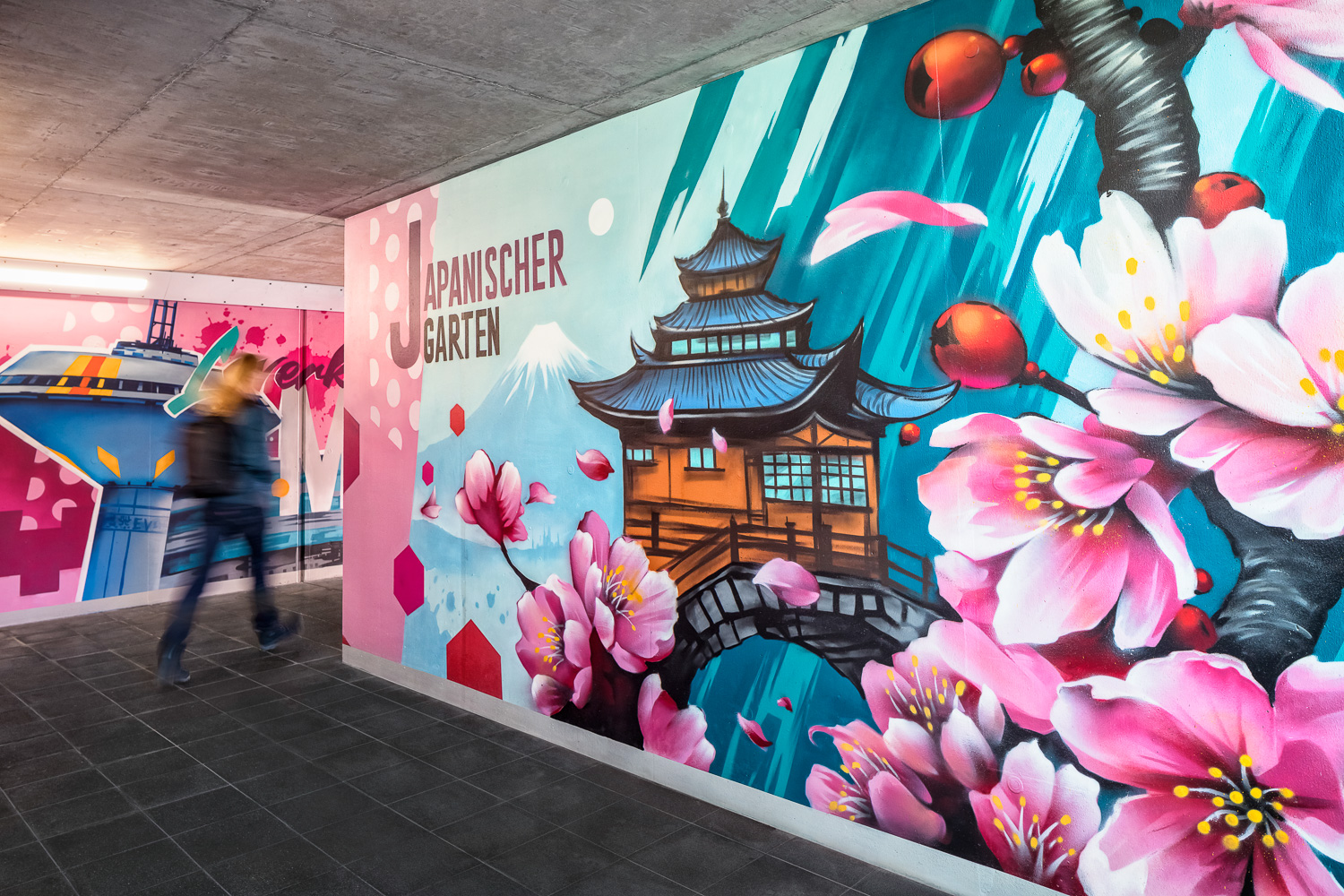 Eine Person geht durch eine Unterführung, die mit farbenfrohen Wandmalereien geschmückt ist, darunter Kirschblüten, ein traditionelles japanisches Gebäude und die Aufschrift "Japanischer Garten" an der Wand.
