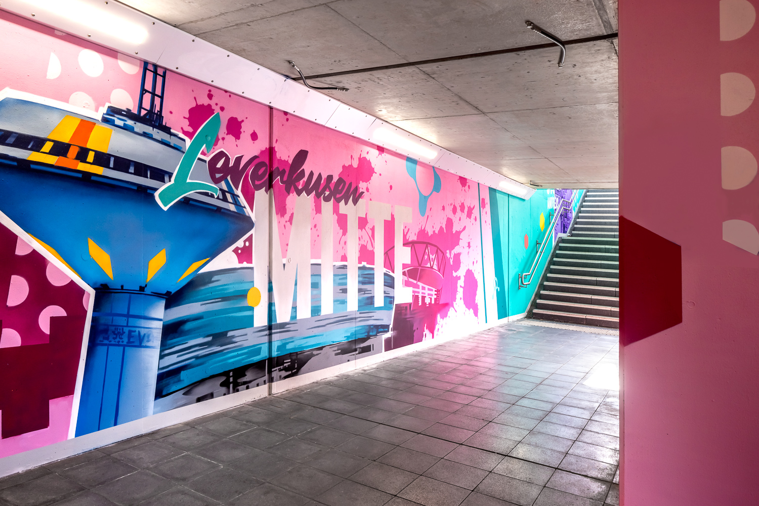 Ein farbenfrohes Wandbild in einer Unterführung zeigt den Schriftzug "Leverkusen MITTE", eine blaue Struktur und abstrakte Formen. Der Tunnel wird durch das Sonnenlicht von den Treppen ins Freie beleuchtet. Wände und Boden sind sauber und modern.