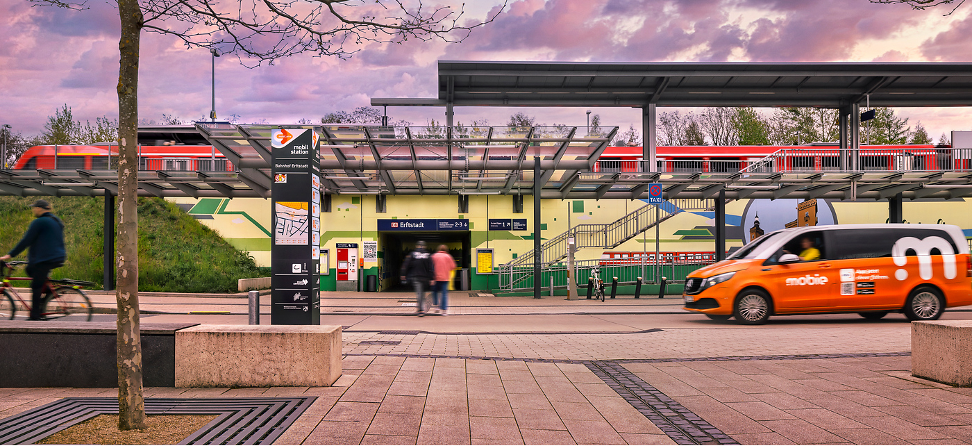 Ein Bahnhofseingang mit einer Treppe, die nach oben führt, ein roter Zug auf Hochbahngleisen, ein Radfahrer und ein orangefarbener Lieferwagen, der im Vordergrund vorbeifährt. Mehrere Personen gehen in der Nähe des Eingangs.