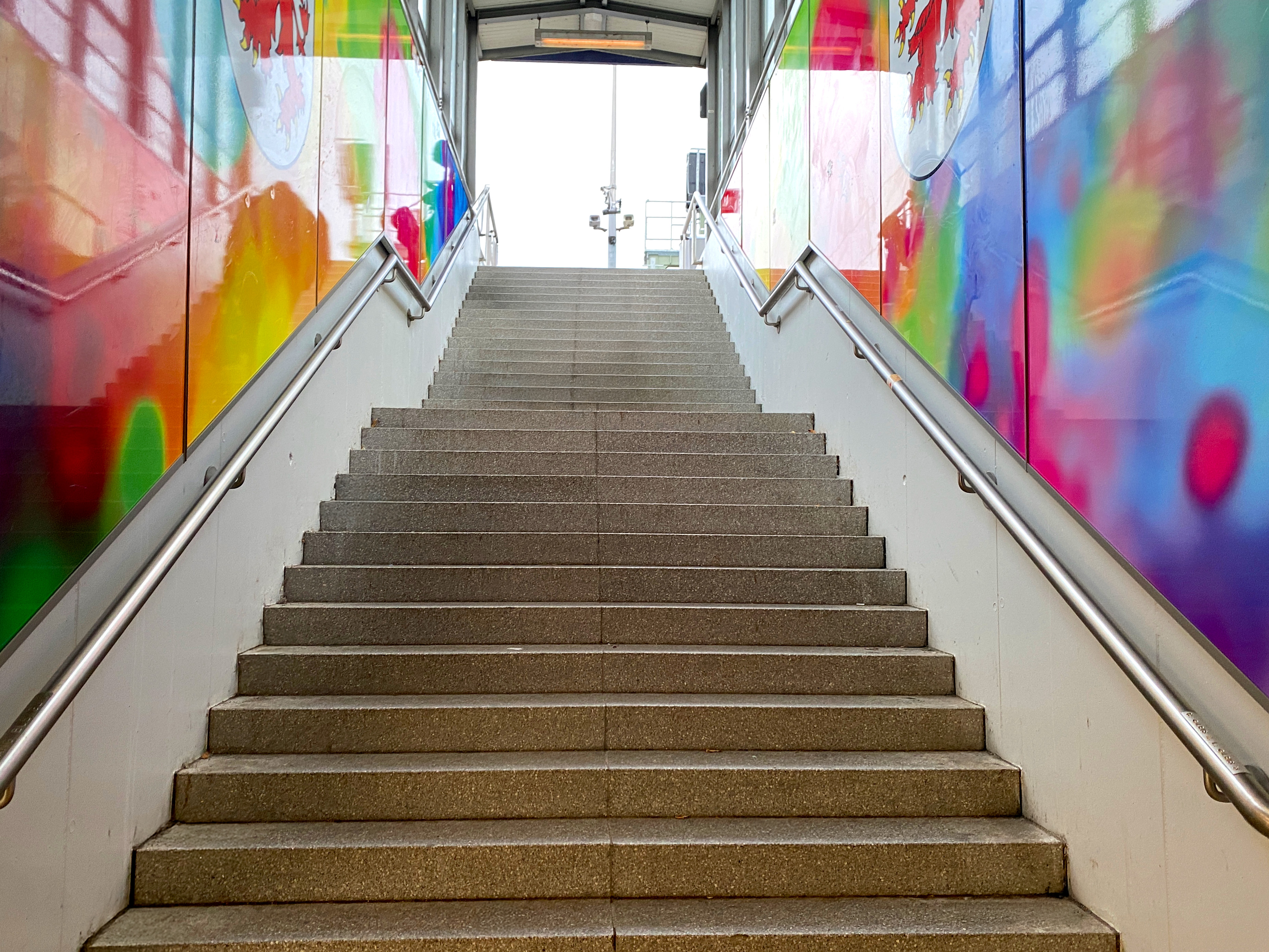 Eine breite, graue Treppe mit Metallgeländern auf beiden Seiten führt zwischen bunten, abstrakten Wandbildern nach oben. Vom oberen Ende der Treppe fällt natürliches Licht ein, das auf einen Ausgang hindeutet.