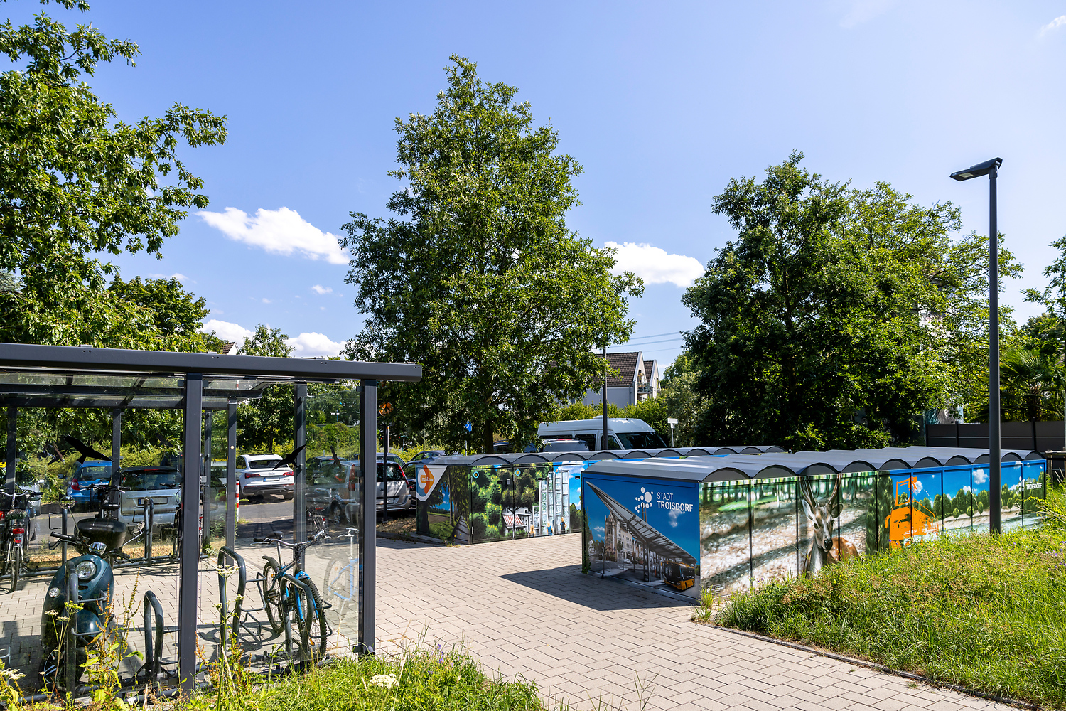 An einem sonnigen Tag steht eine Reihe von überdachten Fahrradabstellplätzen mit farbenfrohen Kunstwerken in der Nähe eines Parkplatzes und von Bäumen, mit einem Glasunterstand für Fahrräder auf der linken Seite und einem gepflasterten Gehweg im Vordergrund.