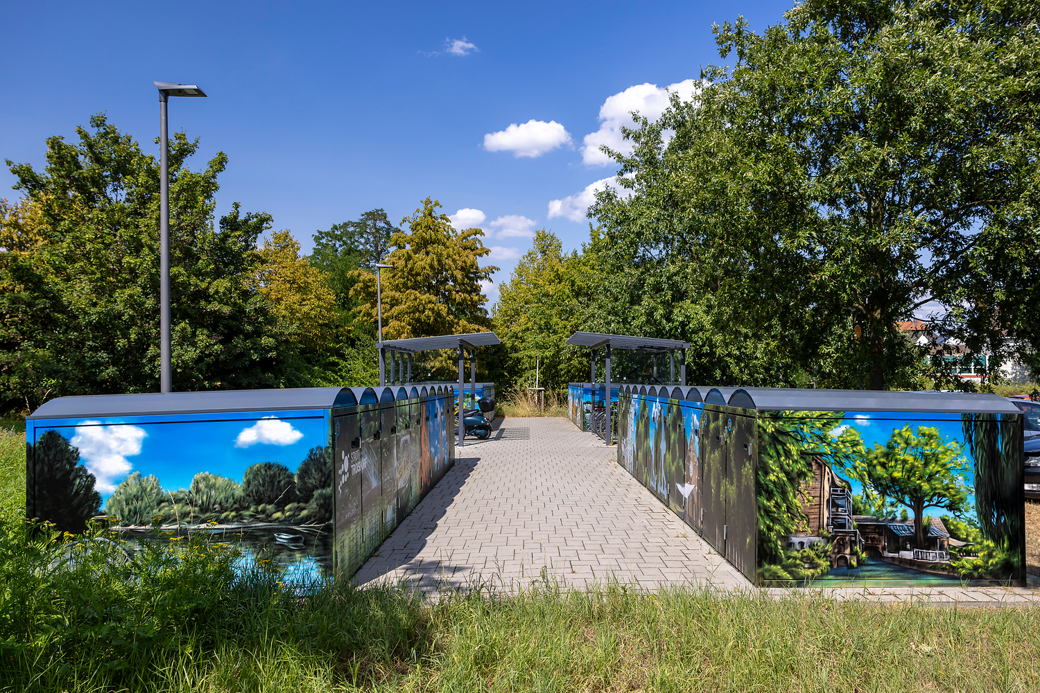 Ein gepflasterter Weg führt zwischen zwei Reihen von überdachten Fahrradabstellanlagen, die jeweils mit Wandmalereien zum Thema Natur dekoriert sind, umgeben von grünen Bäumen unter einem klaren blauen Himmel mit ein paar Wolken.