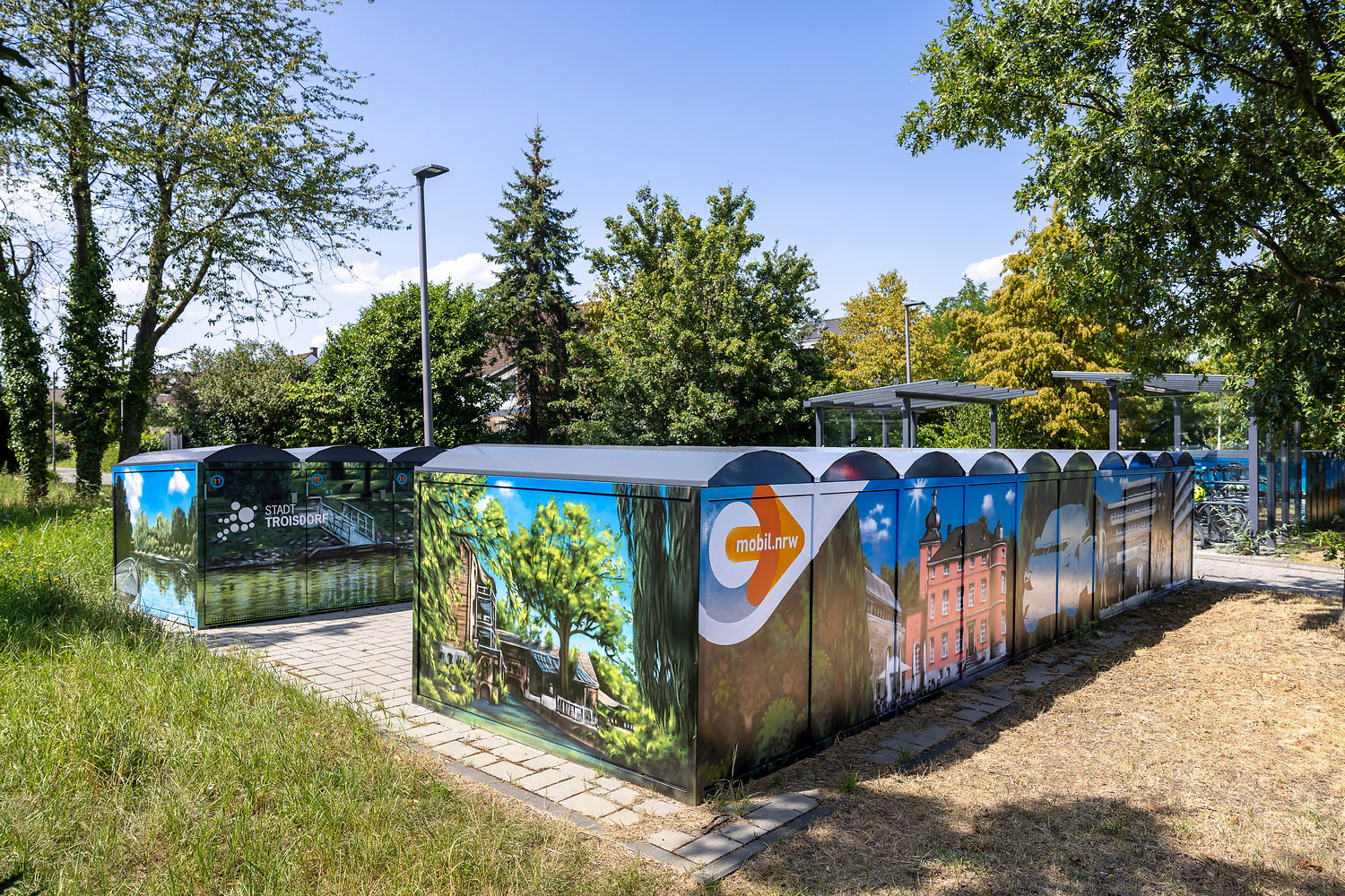 Auf einer gepflasterten Fläche, umgeben von Gras, Bäumen und einigen anderen Strukturen im Hintergrund, steht eine Gruppe von modularen Fahrradabstellanlagen, die mit farbenfrohen Wandbildern aus der Natur und der Stadtlandschaft verziert sind.