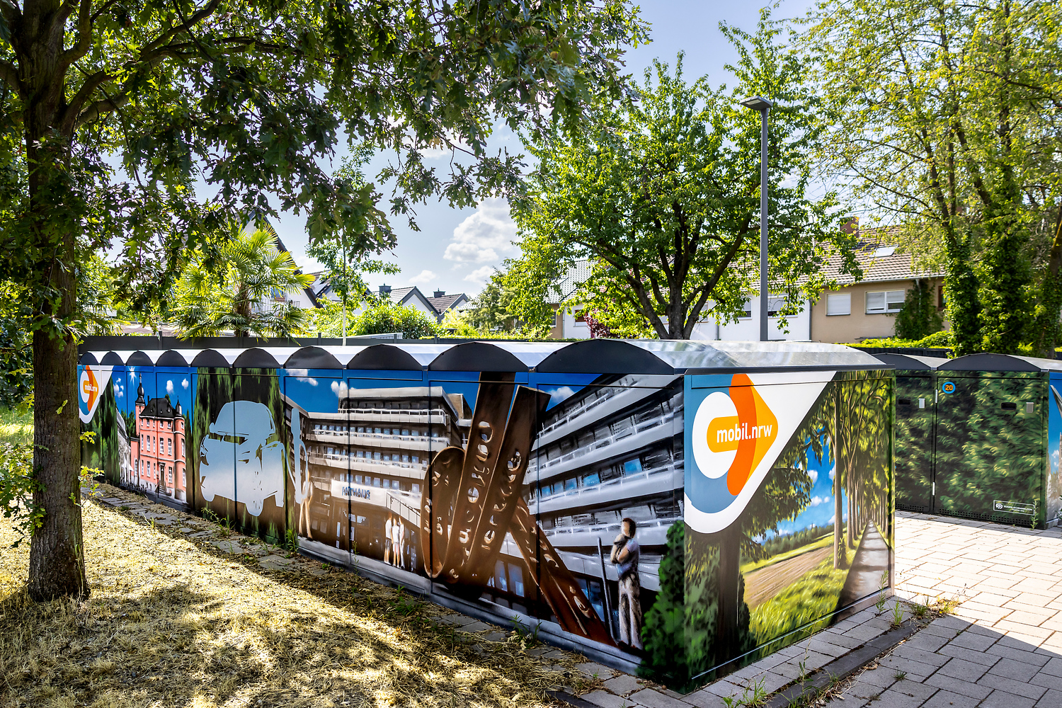 Ein bemalter Stromkasten im Freien zeigt bunte Bilder von Gebäuden, Menschen und einem Baum, auf denen das Logo "mobil.nrw" zu sehen ist. An einem sonnigen Tag ist das Gebiet von Bäumen und Häusern umgeben.