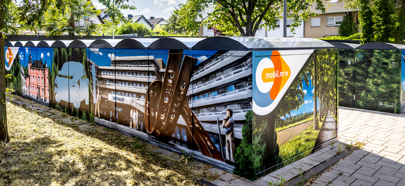 Ein Stromkasten ist mit bunten Wandbildern bedeckt, die Gebäude, einen Zug, eine Brücke, einen von Bäumen gesäumten Weg und eine Person darstellen. Der Kasten trägt das Logo "mobil.nrw" auf mehreren Tafeln. Im Hintergrund sind Häuser und Bäume zu sehen.