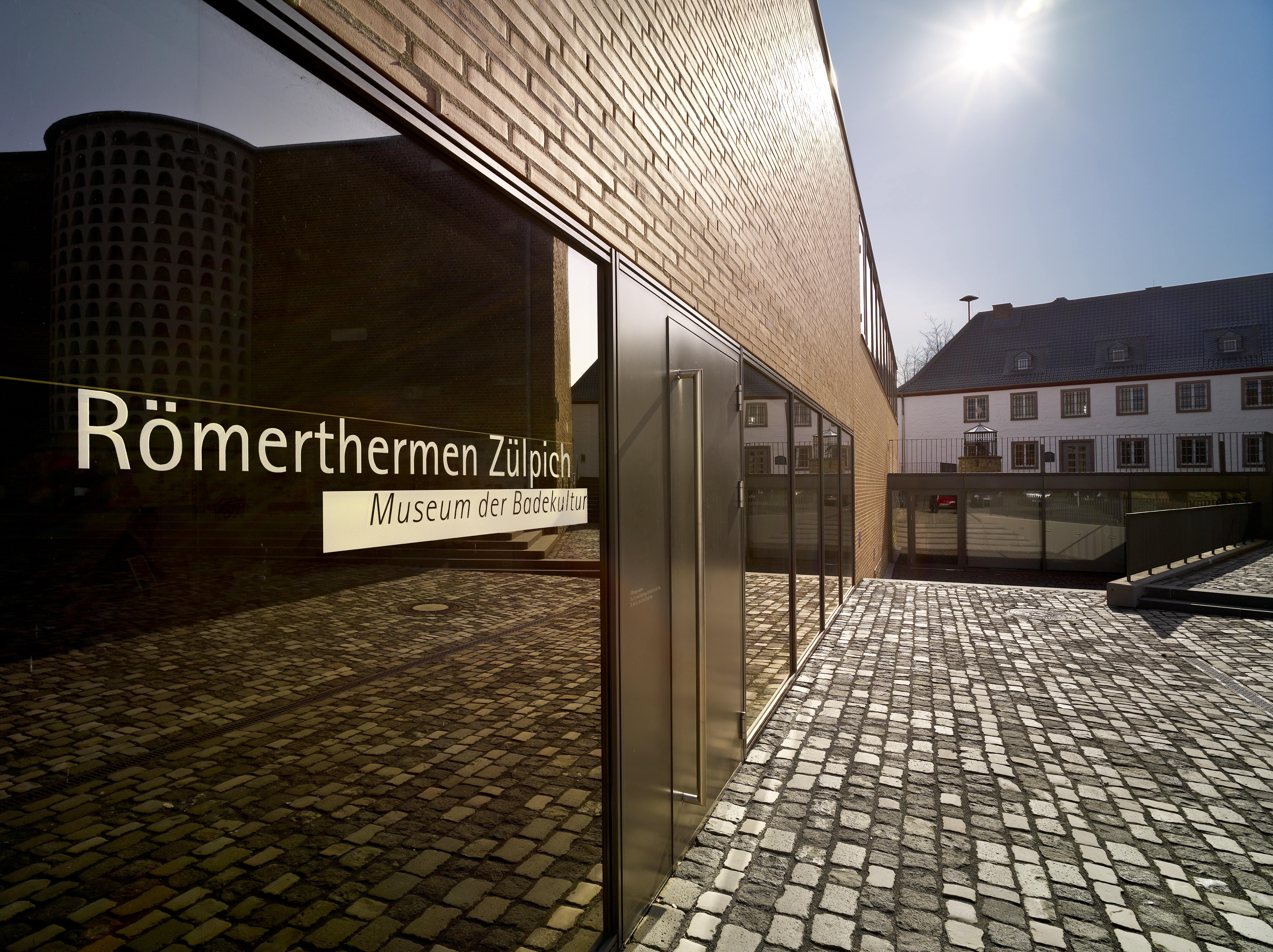 Eingangsbereich des Museums Römerthermen Zülpich mit moderner Glasfassade, die den Namen des Museums spiegelt. Kopfsteinpflaster im Vordergrund, im Hintergrund historische Gebäude unter blauem Himmel.