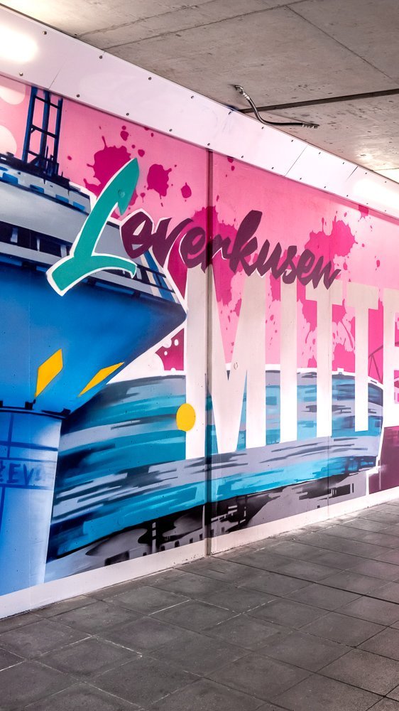 Ein farbenfrohes Wandbild in einer Unterführung zeigt den Schriftzug "Leverkusen MITTE", eine blaue Struktur und abstrakte Formen. Der Tunnel wird durch das Sonnenlicht von den Treppen ins Freie beleuchtet. Wände und Boden sind sauber und modern.