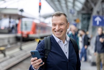 Mann schaut in ein Handy vor einfahrendem Zug im Hintergrund, am Kölner Hauptbahnhof (© go.Rheinland / Smilla Dankert)